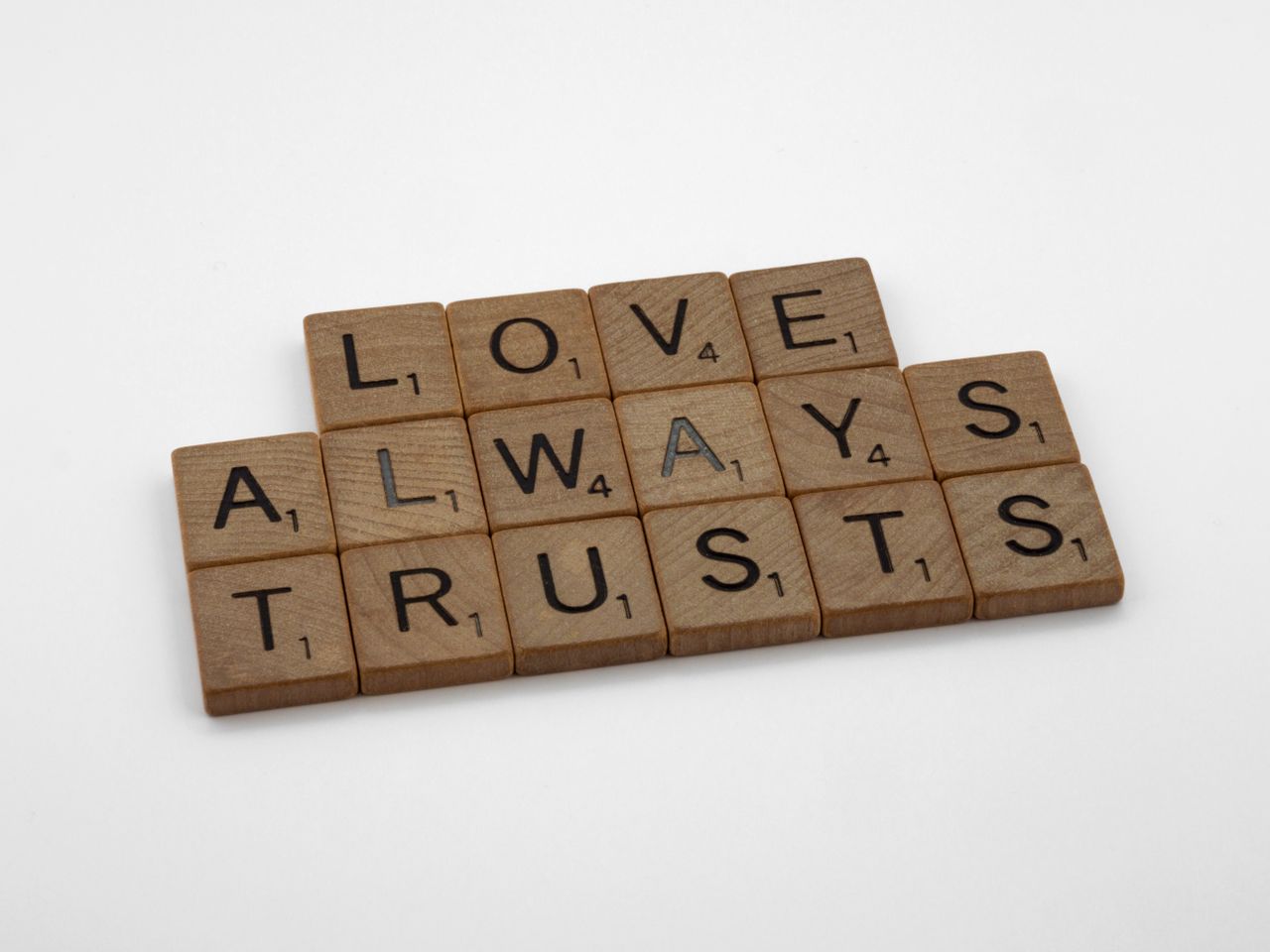 mission-02 Close-up of wooden Scrabble tiles spelling 'Love Always Trusts' on white surface.