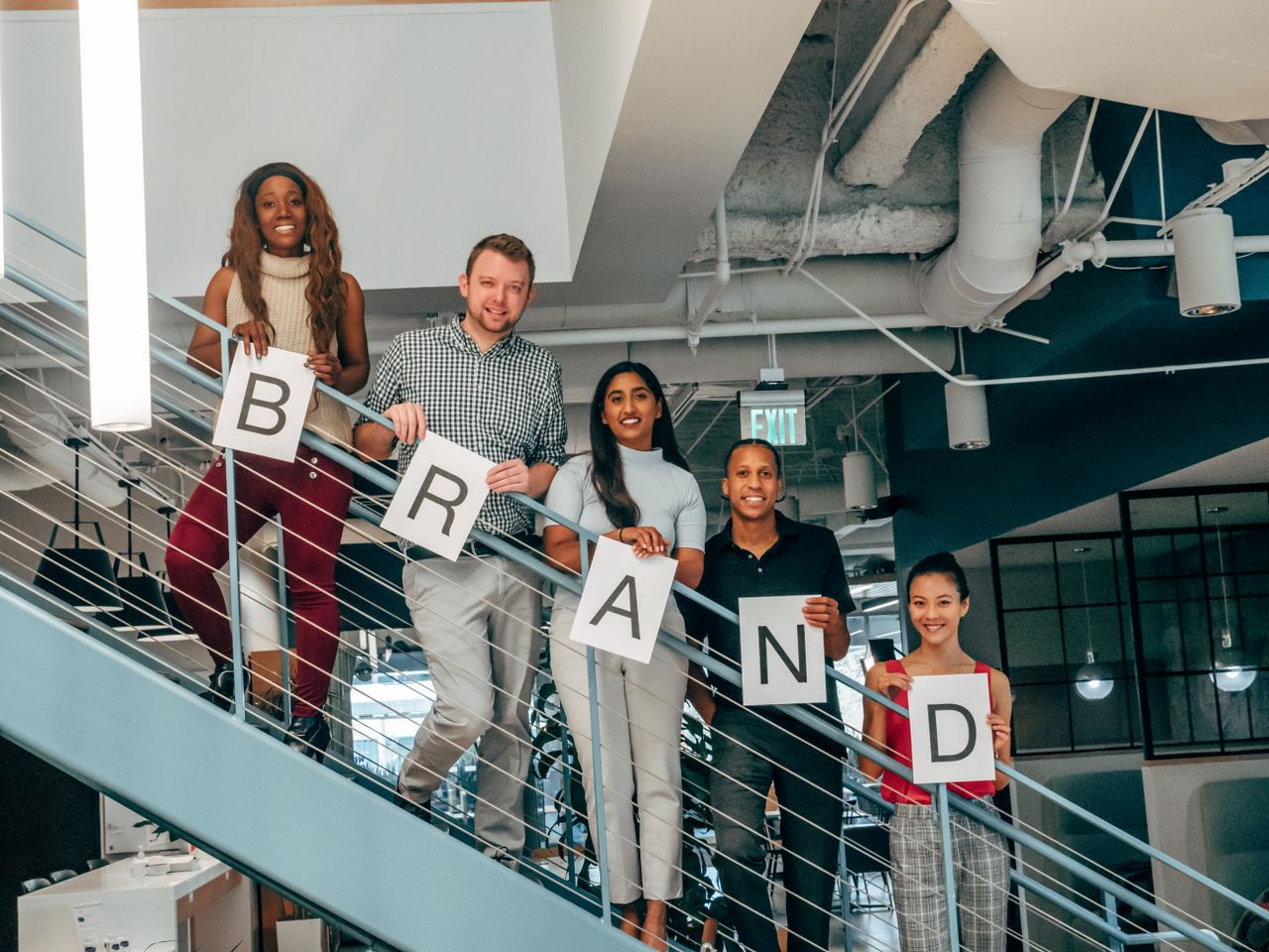 services-05 Diverse group of adults smiling and holding 'BRAND' signs on an office staircase.